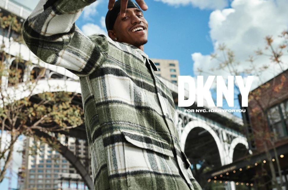 Young man smiles wearing a green and white plaid flannel jacket outdoors with city buildings, blue sky, and DKNY logo in the background.