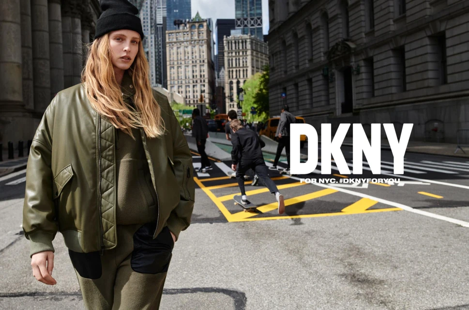 A young adult female poses outdoors in an olive-green bomber jacket and knit cap, with people and city buildings in the background.