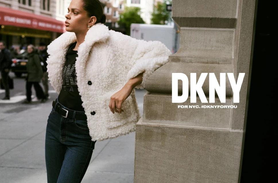 An adult woman with dark hair poses outdoors in an ad for DKNY wearing a sherpa jacket, black shirt, jeans, and belt near a city street.