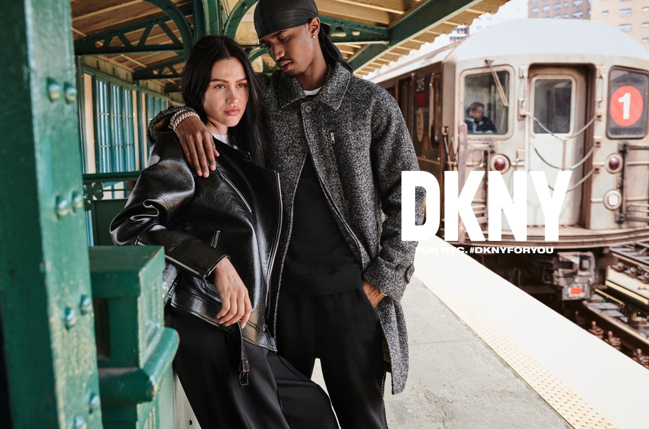 Fashion ad with two models in coats standing on a train platform, train in the background, and DKNY logo overlayed in white.