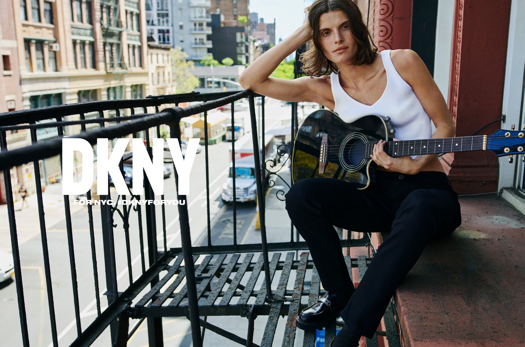 Brunette adult model poses with an acoustic guitar on an outdoor balcony in an urban fashion editorial, wearing a ribbed tank top.