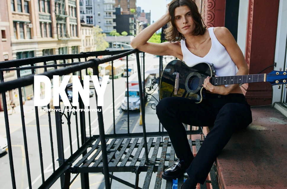Brunette adult model poses with an acoustic guitar on an outdoor balcony in an urban fashion editorial, wearing a ribbed tank top.