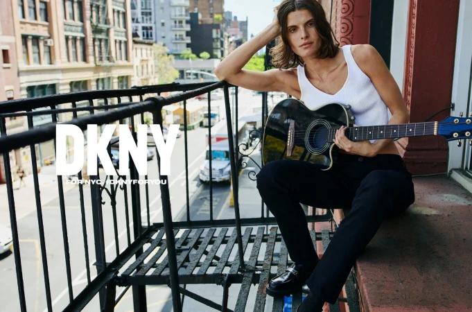 Brunette adult model poses with an acoustic guitar on an outdoor balcony in an urban fashion editorial, wearing a ribbed tank top.
