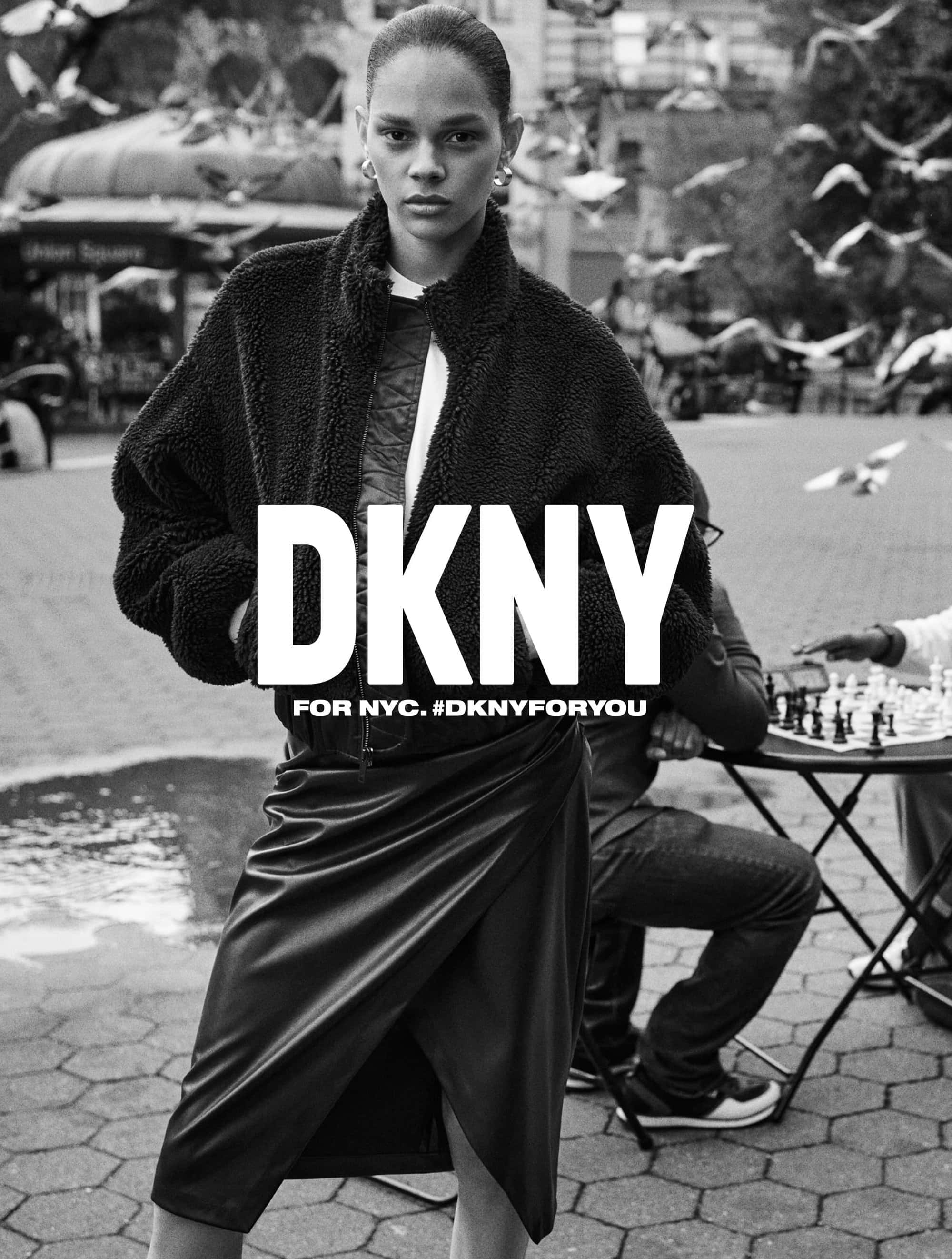 Black-and-white fashion shot in an urban square, featuring a stylish woman in an outfit and a man playing chess in the backdrop.