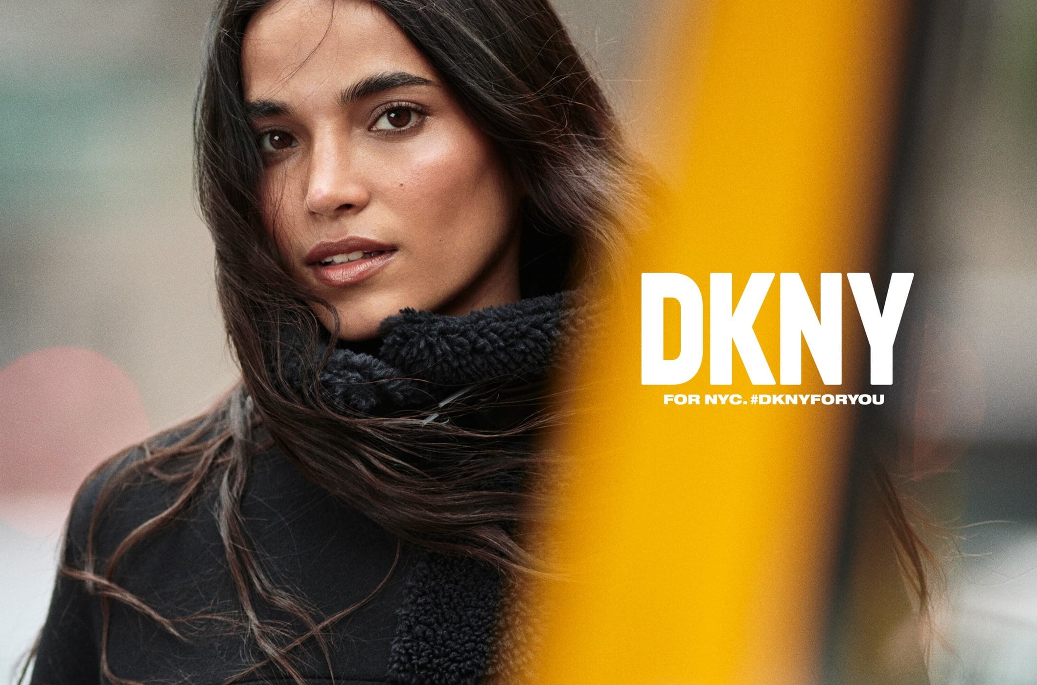 Brunette woman in a close-up fashion shot. She wears a black jacket with fur trim. A yellow column and text add graphic elements.
