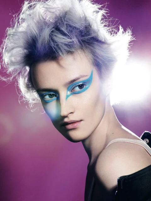 Close-up portrait of a young adult female with theatrical blue eye makeup, white and purple hair, and magenta studio background.