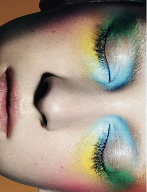 Close-up studio shot of a woman's face with colorful eyeshadow and closed eyes, set against a warm, orange background.