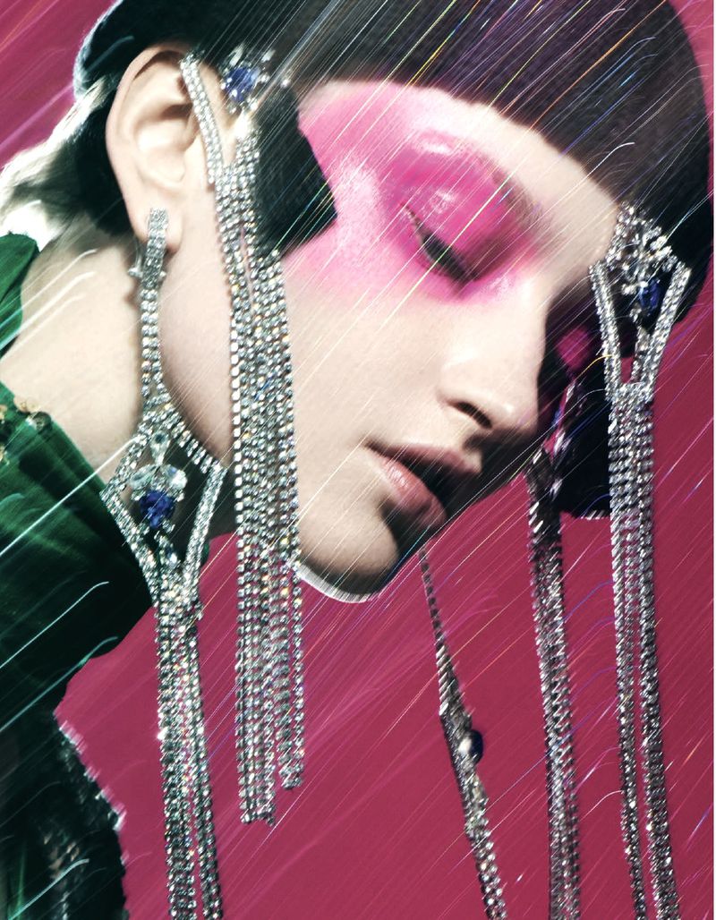 Close-up of a woman with pink eyeshadow and jeweled headpiece and earrings against a pink backdrop in a beauty and fashion portrait.