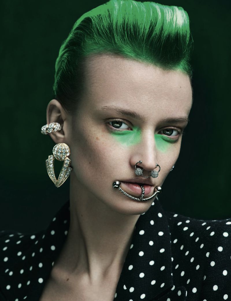 Close-up portrait of a woman with green hair, heavy jewelry, a polka dot blazer, and green makeup against a dark backdrop.