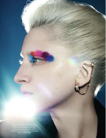 Portrait of a woman with creative makeup featuring colorful eyeshadow, gold earrings, and platinum blonde hair against a blue backdrop.