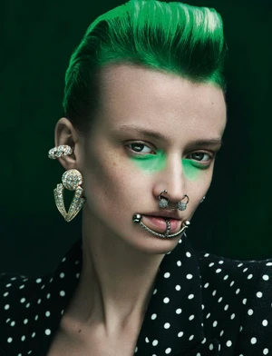 Model with vibrant green hair and matching eye makeup wearing elaborate facial piercings and a polka dot jacket against a dark green background