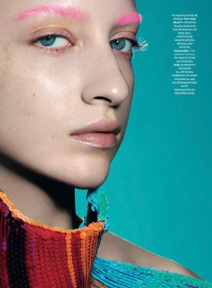 Close-up portrait of a model with bright pink eyebrows and colorful sequined clothing against a vibrant cyan background.