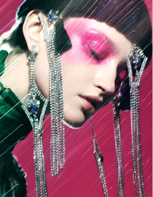 Close-up of a model with dramatic pink makeup and elaborate cascading rhinestone earrings against a magenta background.