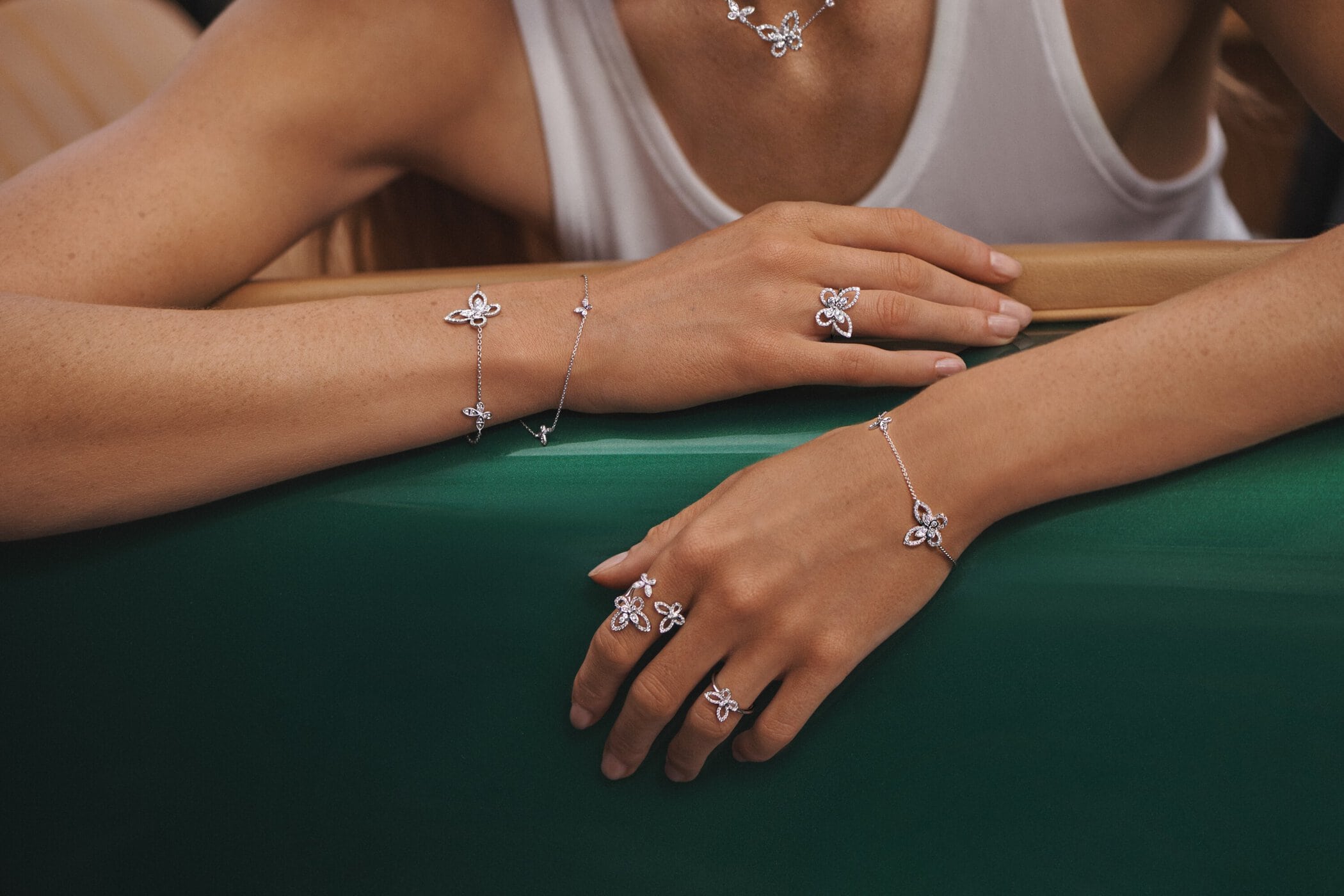 Close-up of a woman's hands and arms adorned with diamond butterfly rings, bracelets and necklace against a green backdrop.