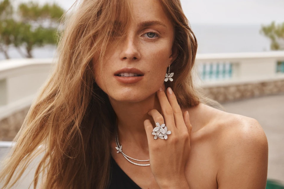 Close-up of a blonde woman wearing butterfly jewelry including a ring, necklace, and earrings, with a blurred outdoor background.