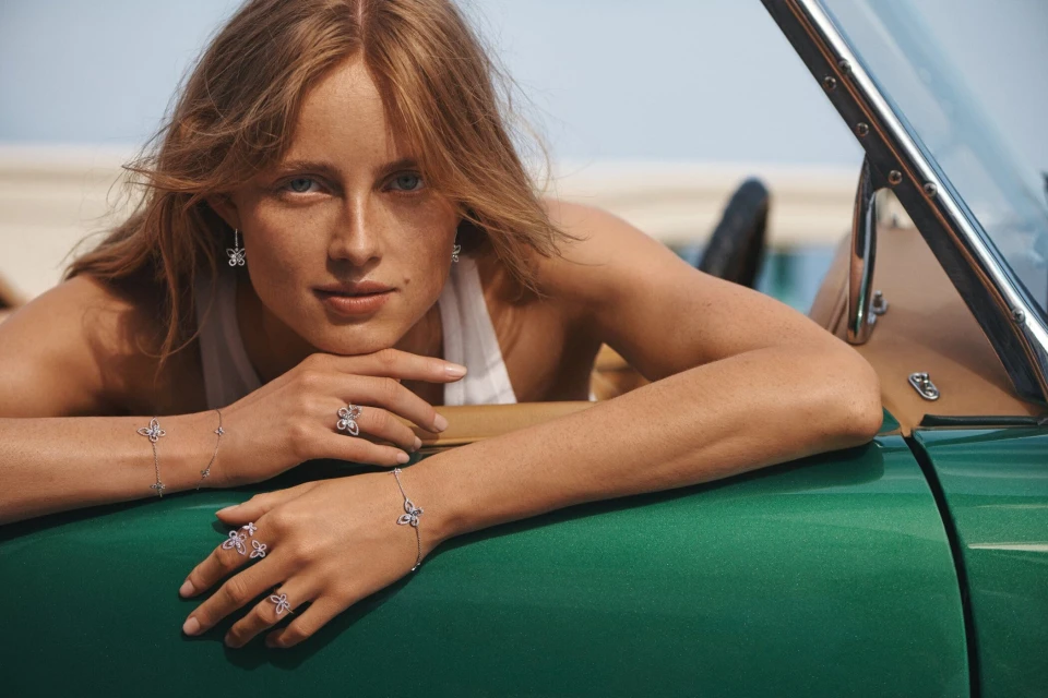 Woman poses with jewelry in a green convertible car on a beach with light blue sky, showcasing butterfly-themed rings, earrings, and bracelets.
