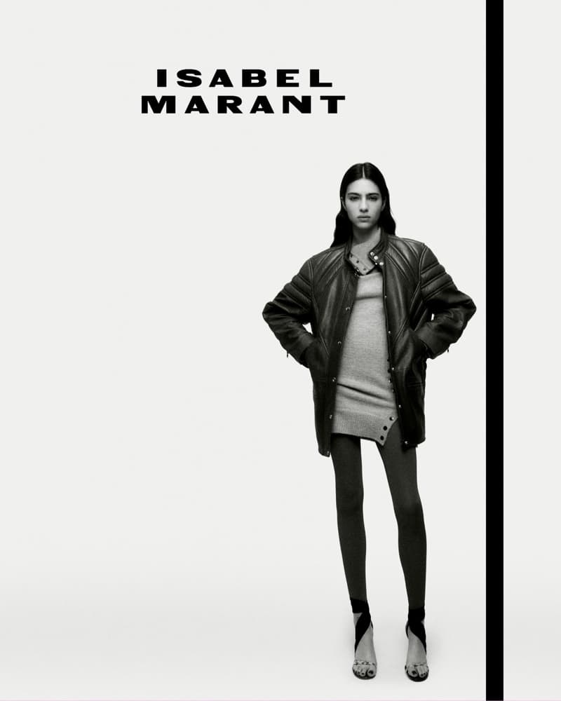 Black and white fashion shot of a female model wearing a jacket and tights with hands on hips against a white backdrop.