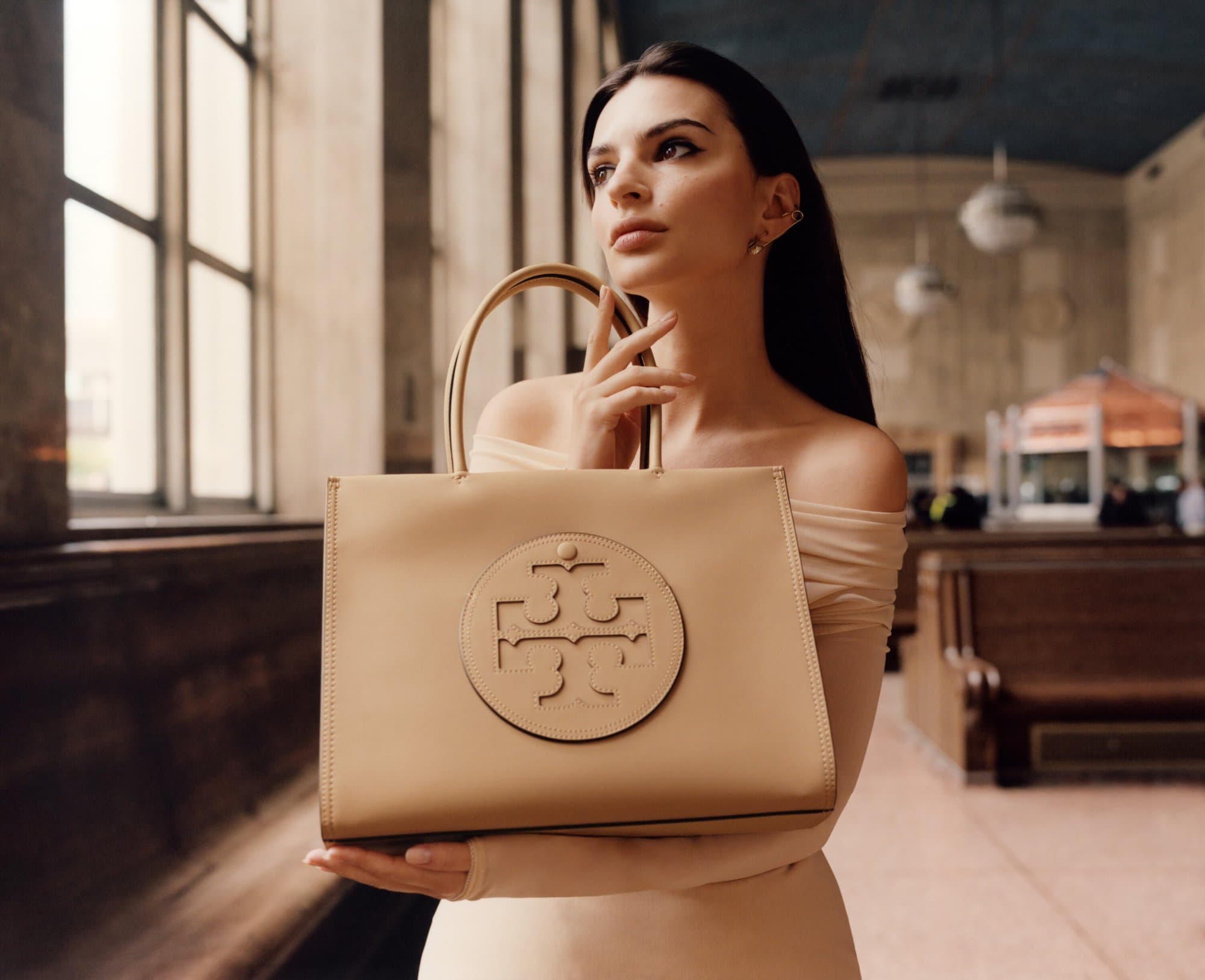 Woman poses with tan leather Tory Burch tote bag, indoor location with window light and neutral tones, fashion advertising. 