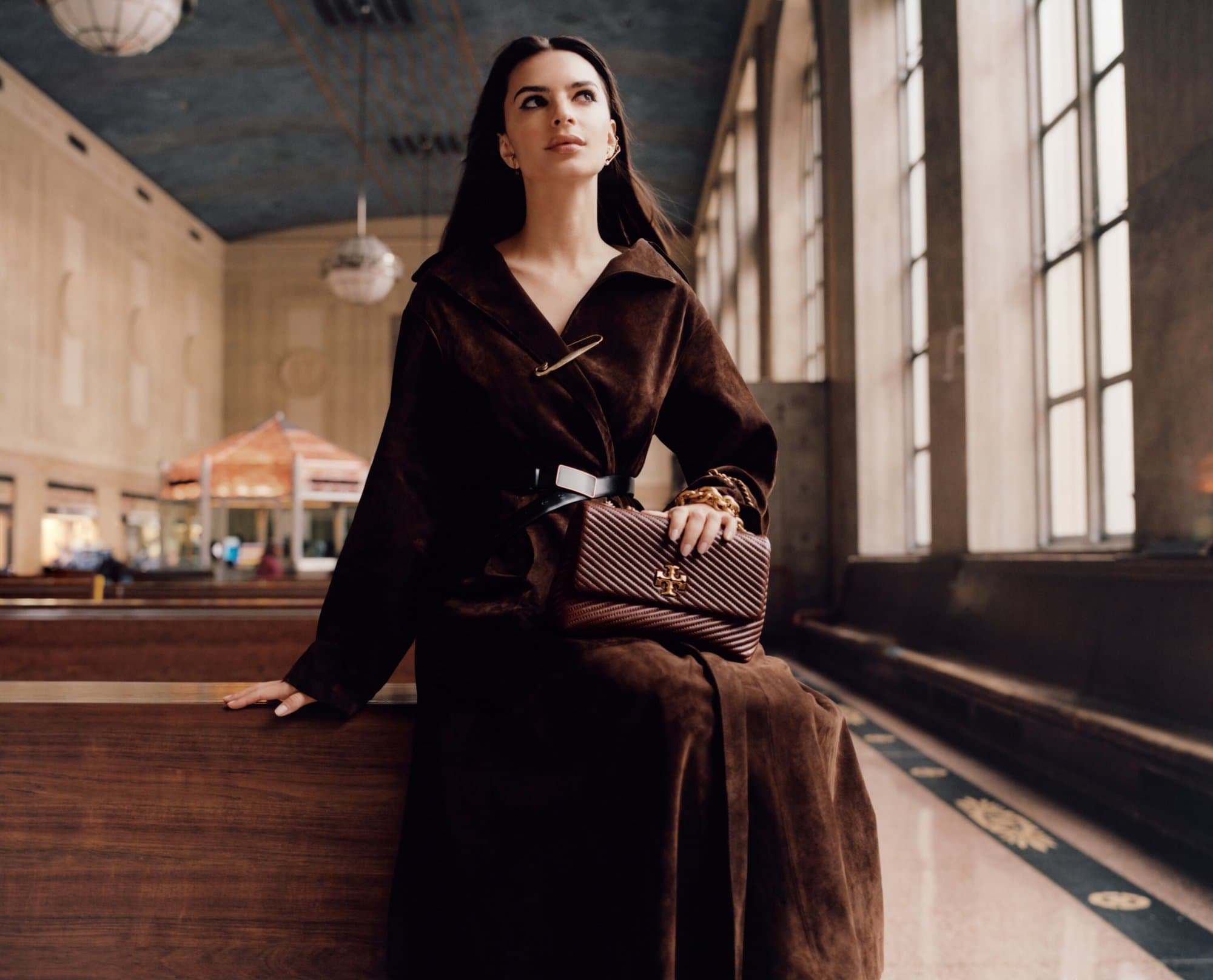 A woman in a brown coat sits in a train station, holding a purse, looking up toward the skylight with large windows, and decorative features.