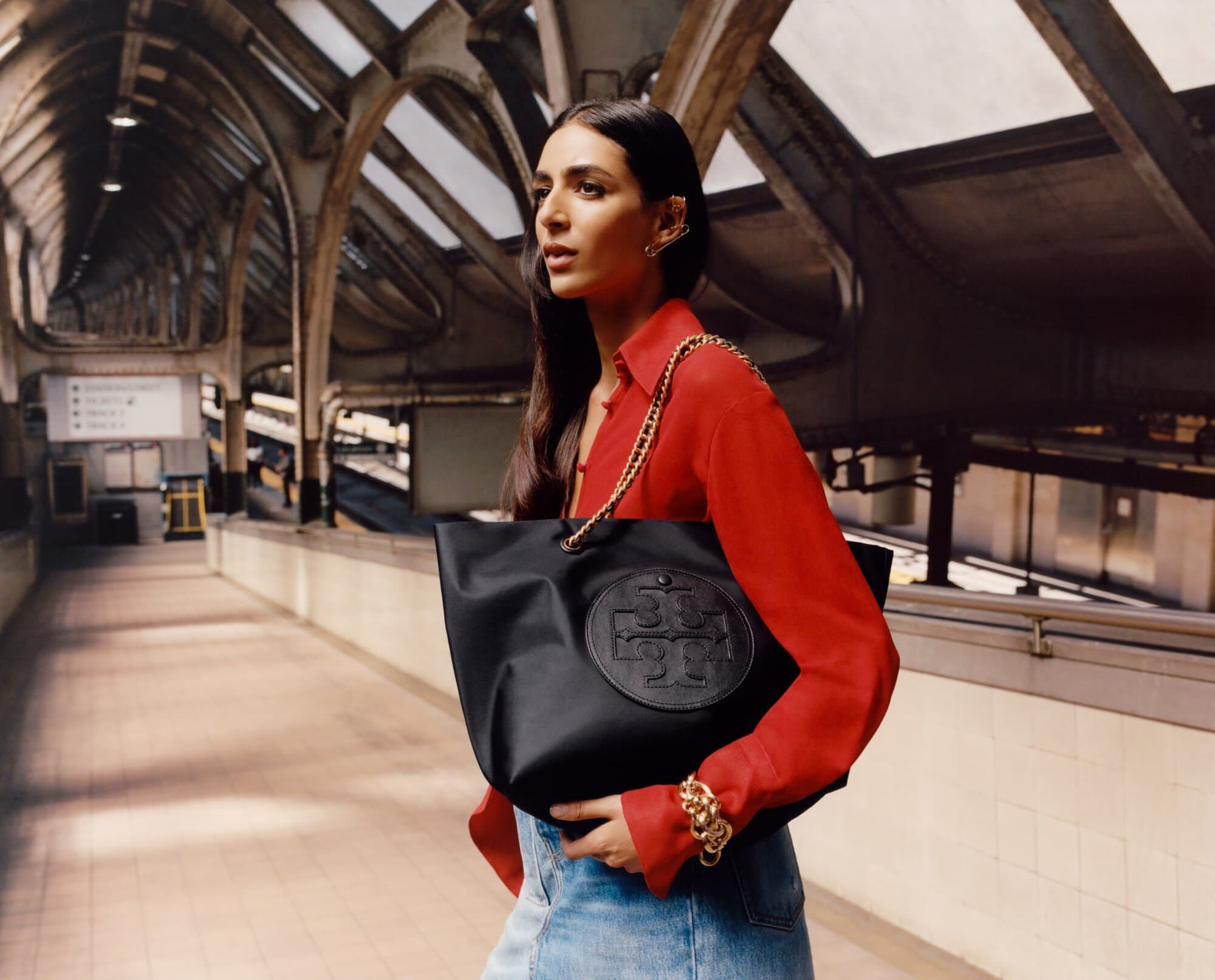 A woman in a red blouse and jeans carries a black tote bag with a logo inside a subway station, creating a fashionable and urban scene.