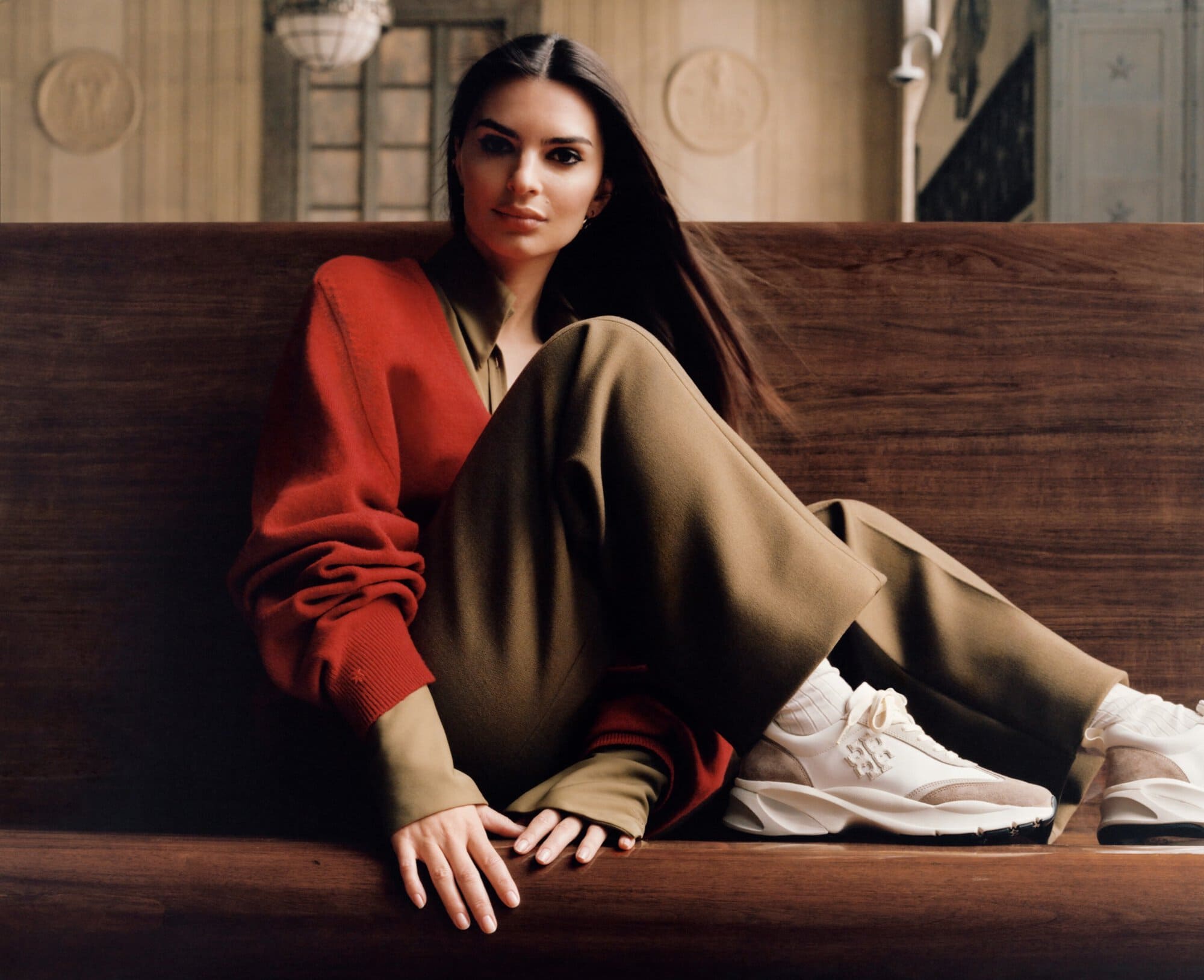 A brunette woman sits on a dark wooden bench wearing tan pants, white sneakers, and a red sweater with a light-brown button-down shirt.