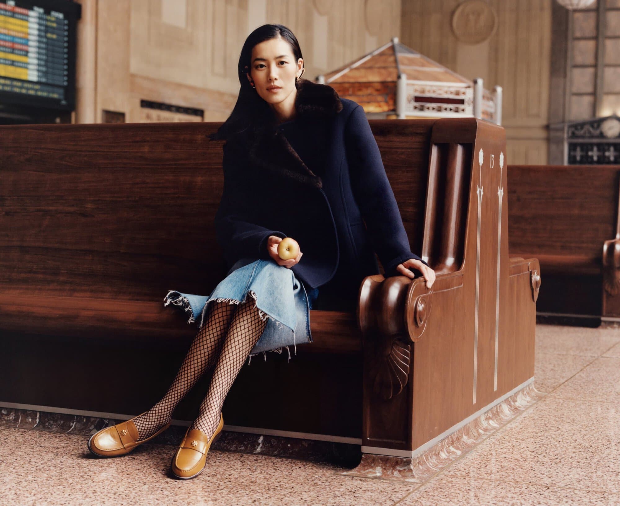 An Asian woman wearing a dark coat, denim skirt, fishnet stockings, and loafers sits on a wooden bench holding an apple inside a building.