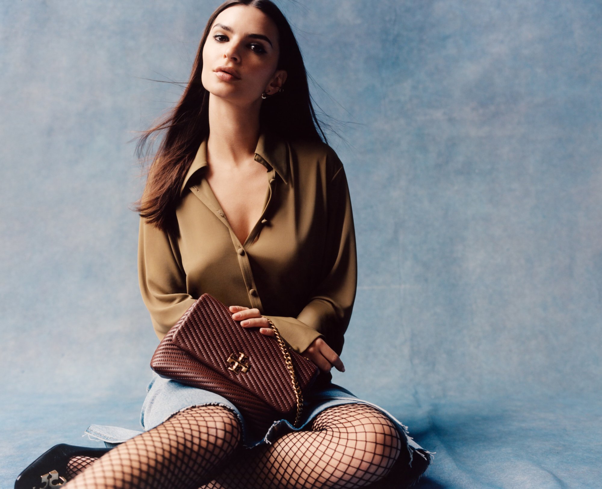 Brunette woman poses in olive green shirt, blue denim skirt, black fishnet stockings, and flats, holding burgundy purse with gold chain.