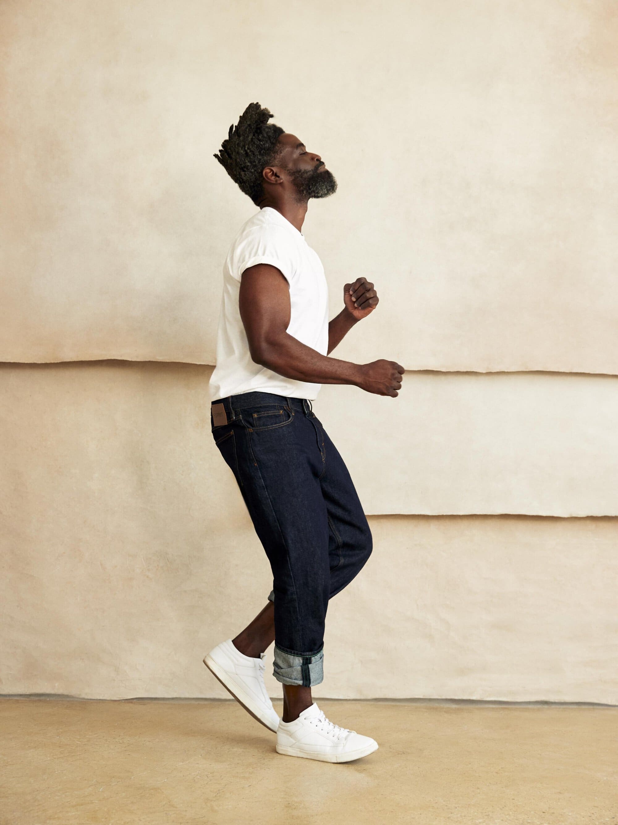 Full-length portrait of a black man in jeans and a white t-shirt standing against a neutral textured wall. He is looking up and smiling.