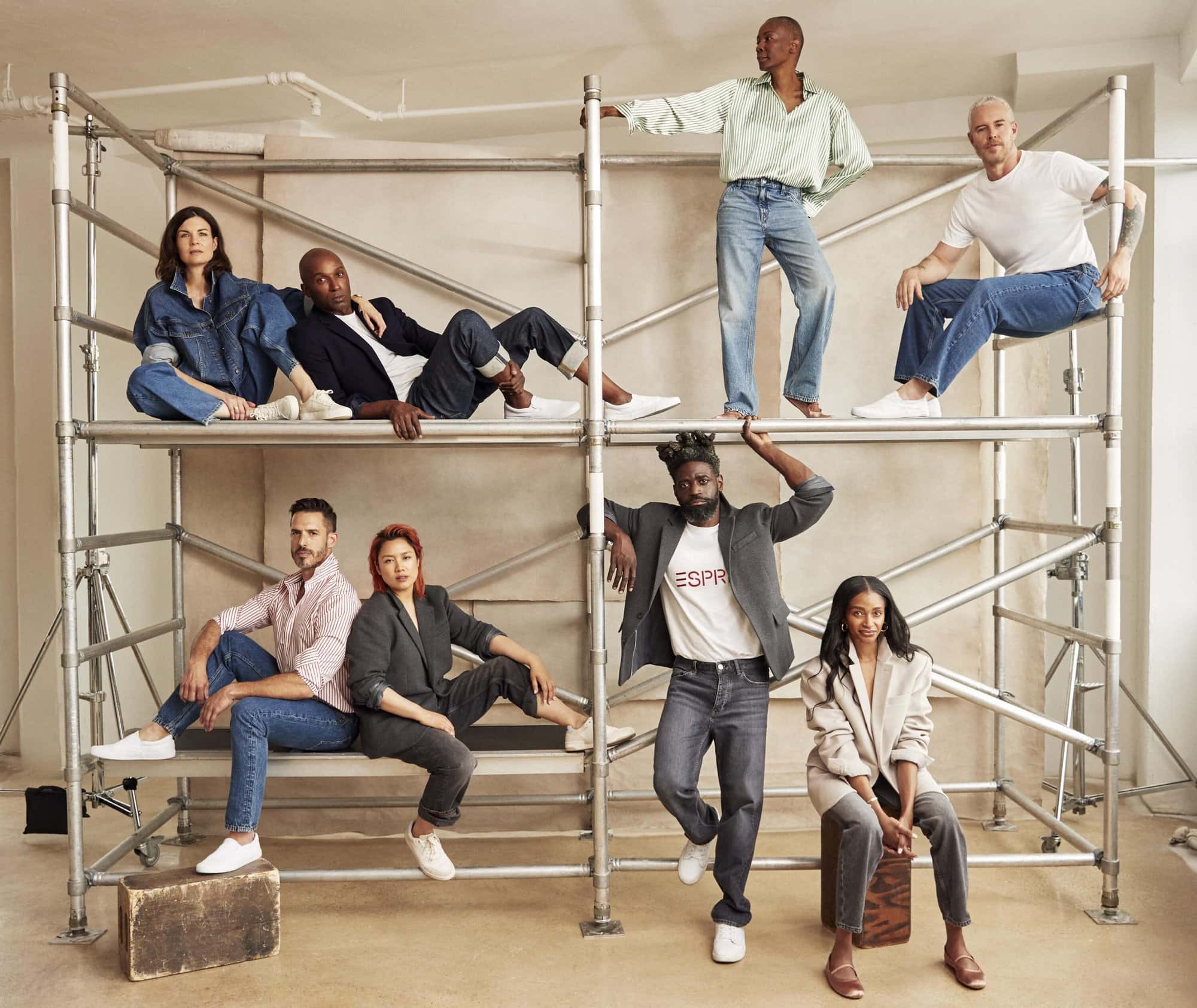 Group of seven diverse adults in a photo studio, posing casually on and around a metal scaffolding structure, wearing casual clothing and white sneakers.