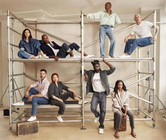Group of seven diverse adults in a photo studio, posing casually on and around a metal scaffolding structure, wearing casual clothing and white sneakers.
