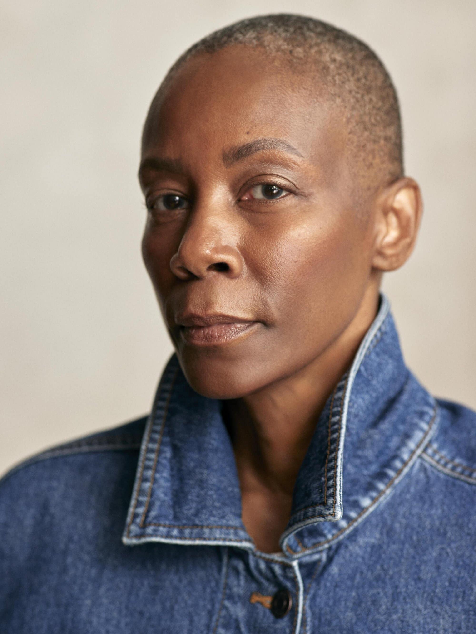 Close-up portrait of an adult woman with shaved hair, wearing a blue denim jacket, against a neutral background in an indoor setting.