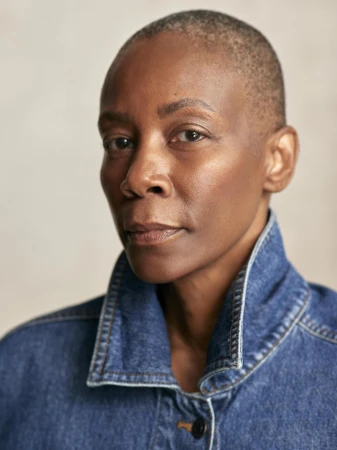 Close-up portrait of an adult woman with shaved hair, wearing a blue denim jacket, against a neutral background in an indoor setting.