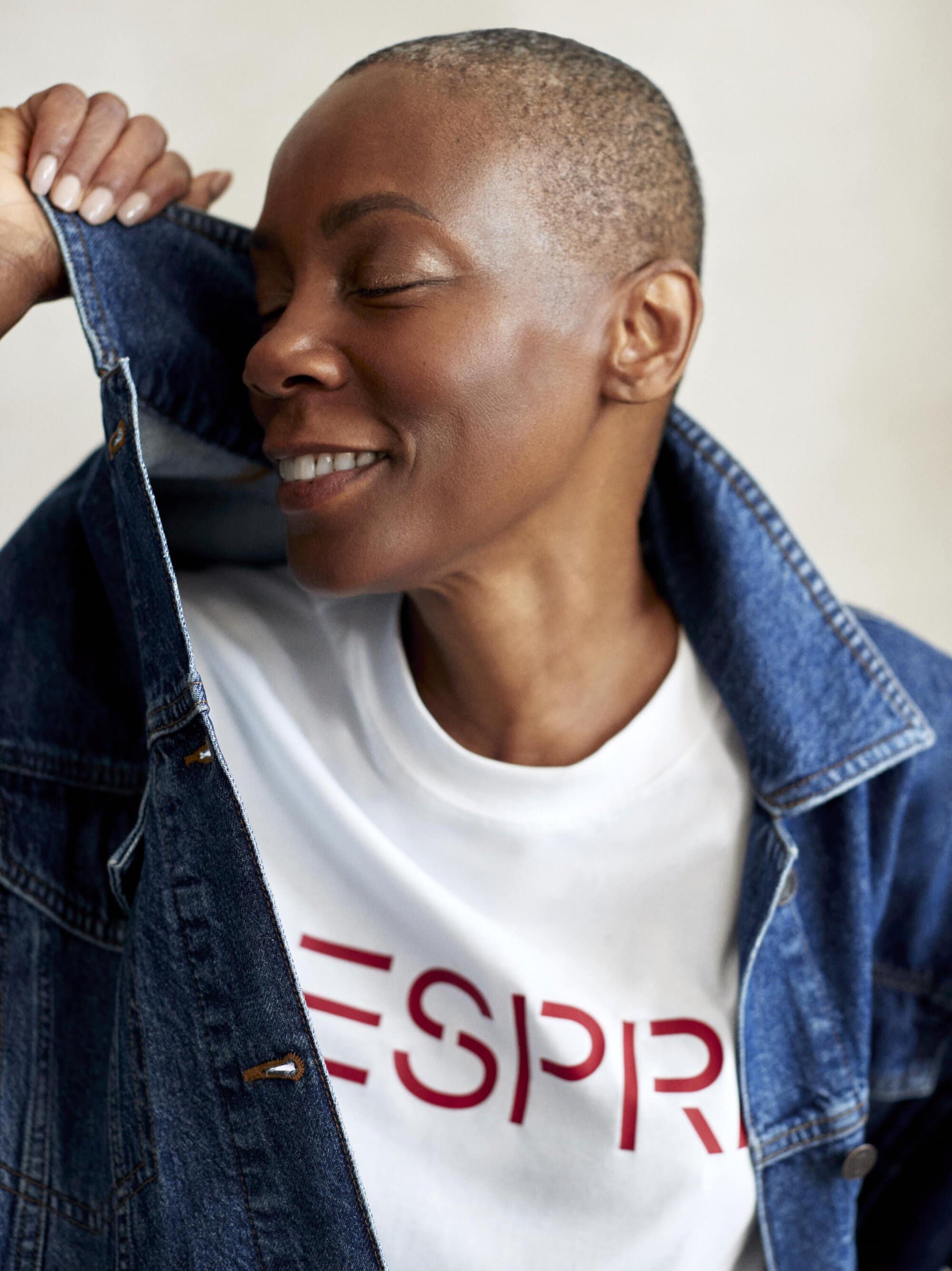 Smiling woman in a denim jacket and white t-shirt with closed eyes. The woman has a shaved head. The background is blurred.