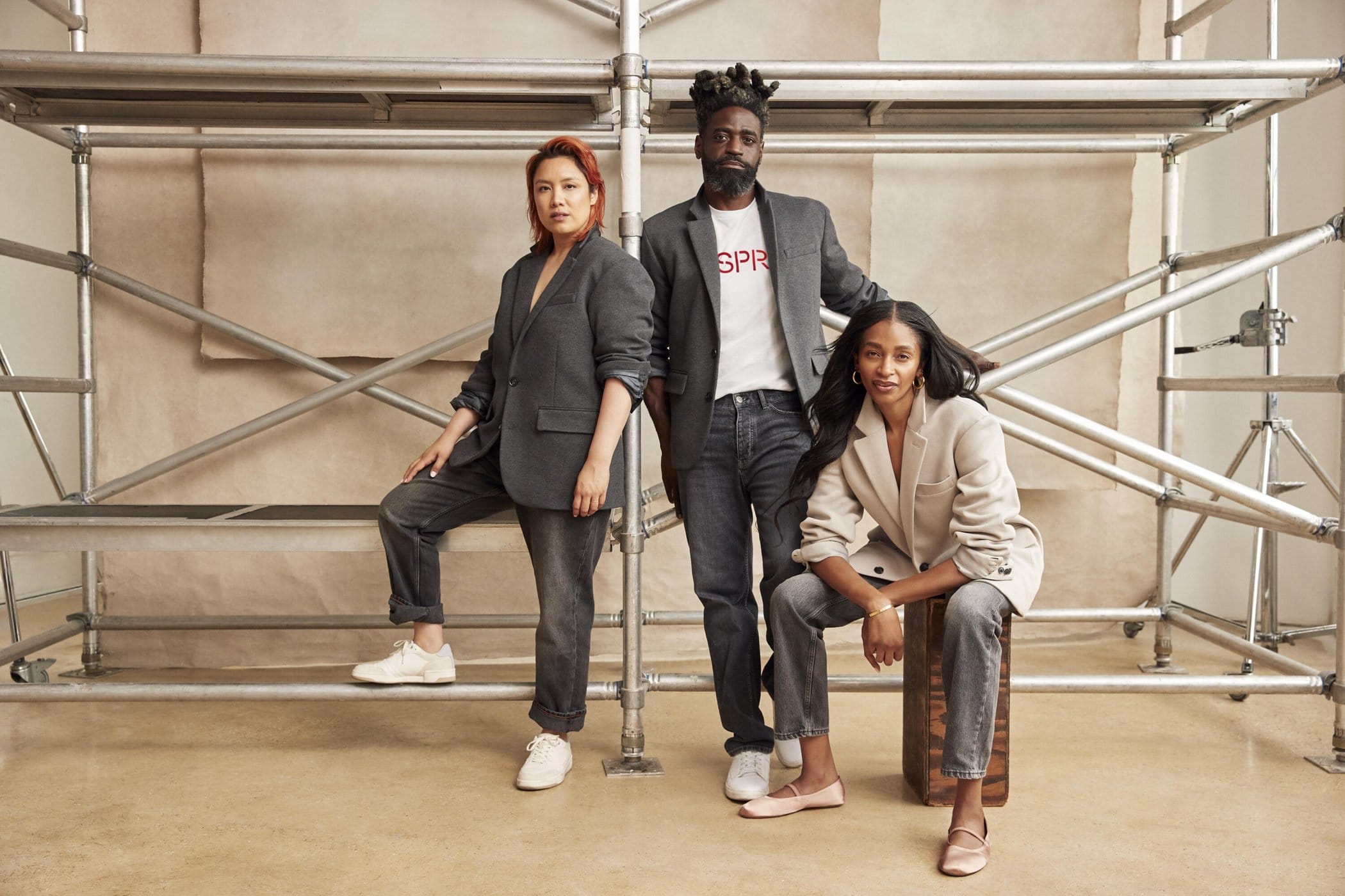 Three adult models—an Asian woman, a Black man, and a Black woman—pose in a photo studio with a scaffolding structure, wearing grey and beige blazers.