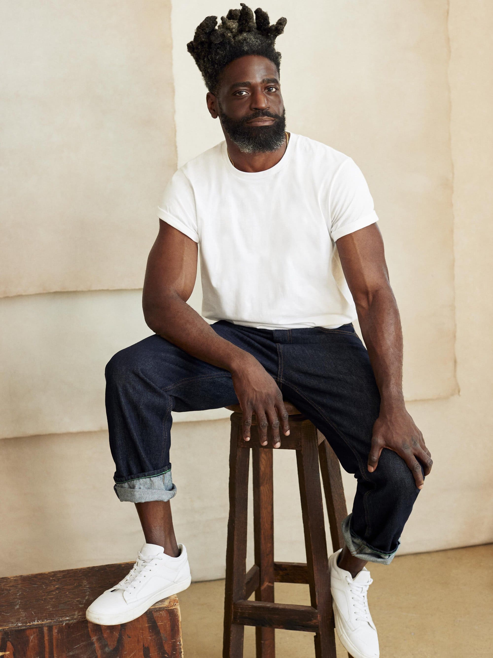 Stylish Black male model poses sitting on a wooden stool in casual attire against a neutral backdrop in a studio setting.