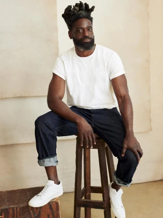 Stylish Black male model poses sitting on a wooden stool in casual attire against a neutral backdrop in a studio setting.