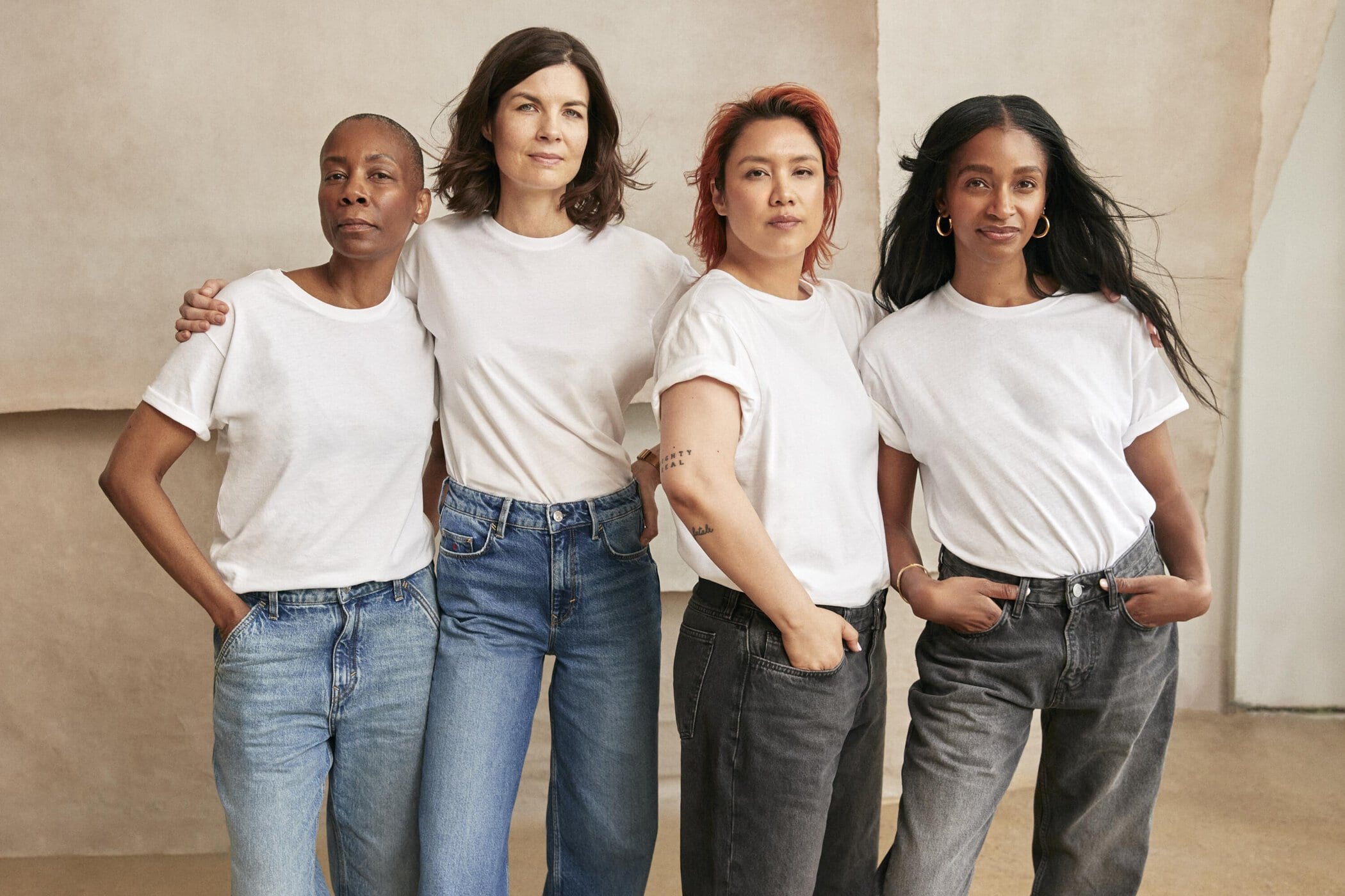 Diverse group of four adult women in casual white t-shirts and jeans, standing close together, indoors with neutral backdrop in a portrait.