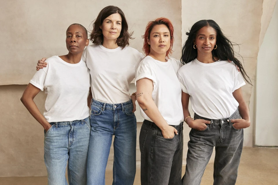 Diverse group of four adult women in casual white t-shirts and jeans, standing close together, indoors with neutral backdrop in a portrait.