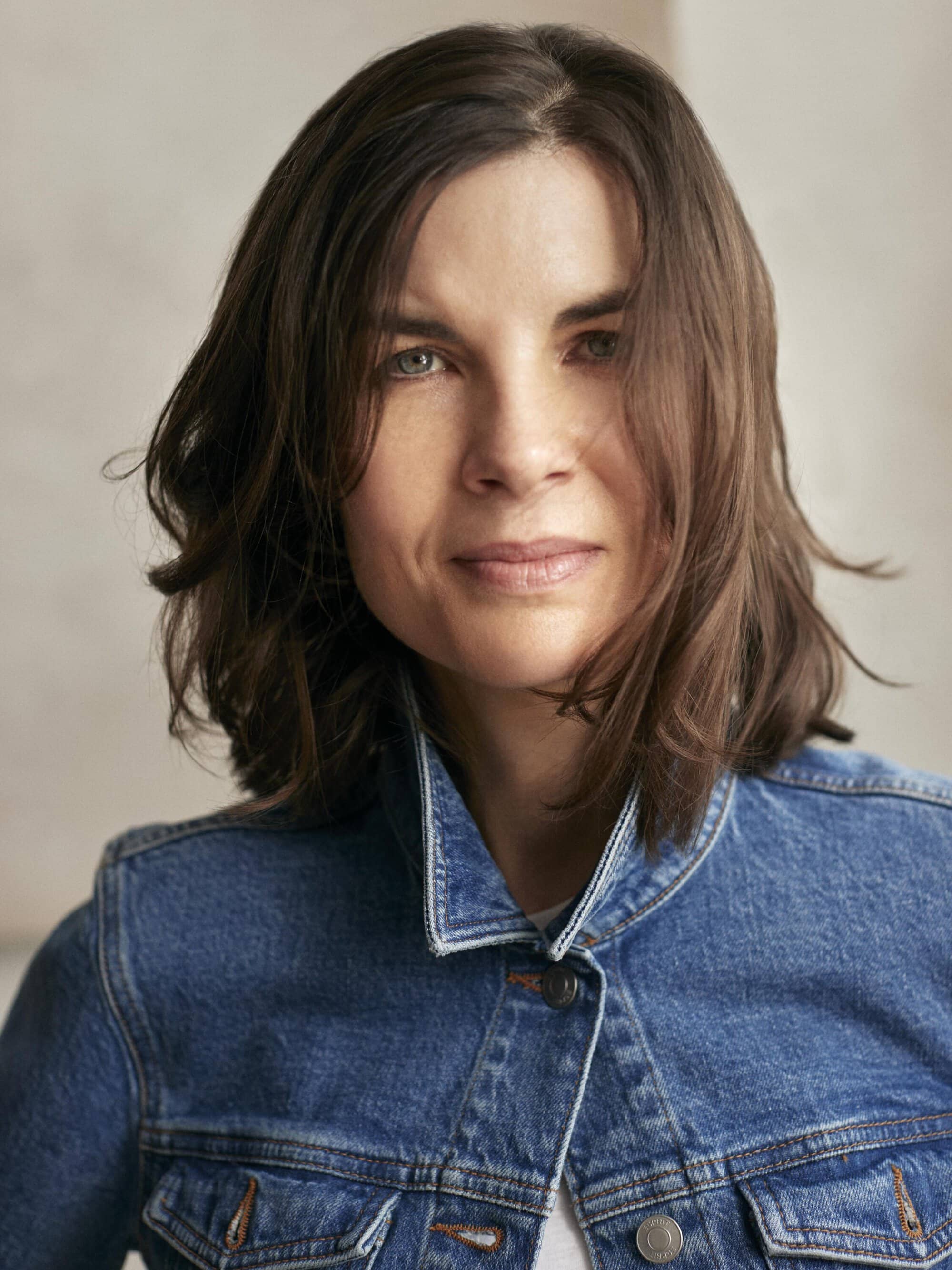 Close-up portrait of an adult woman with medium-length brown hair wearing a blue denim jacket against a neutral background.