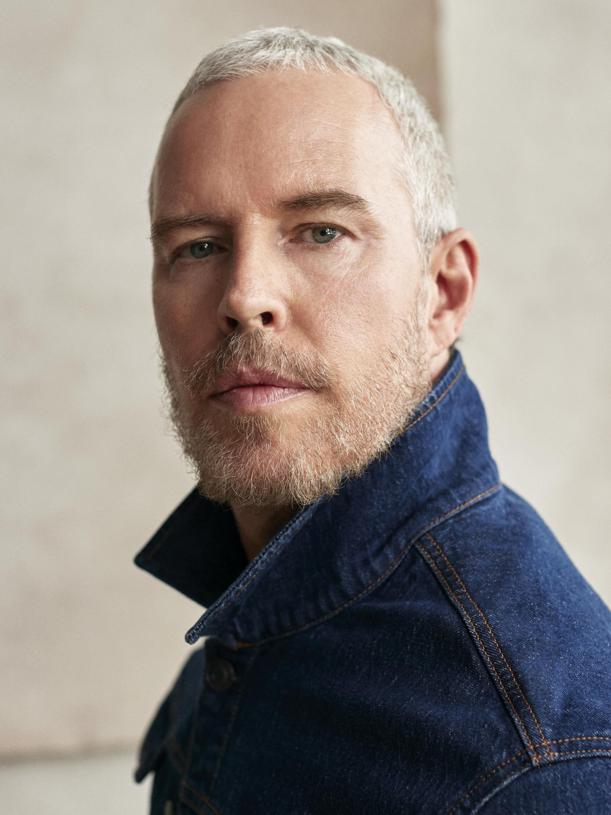 Close-up portrait of a mature adult male with short grey hair and a denim jacket against a neutral background, showcasing a clean and modern look.