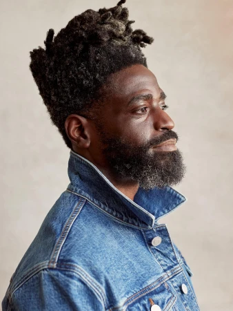 Profile portrait of a Black man with a textured afro hairstyle and full beard, wearing a classic blue denim jacket against a plain background.