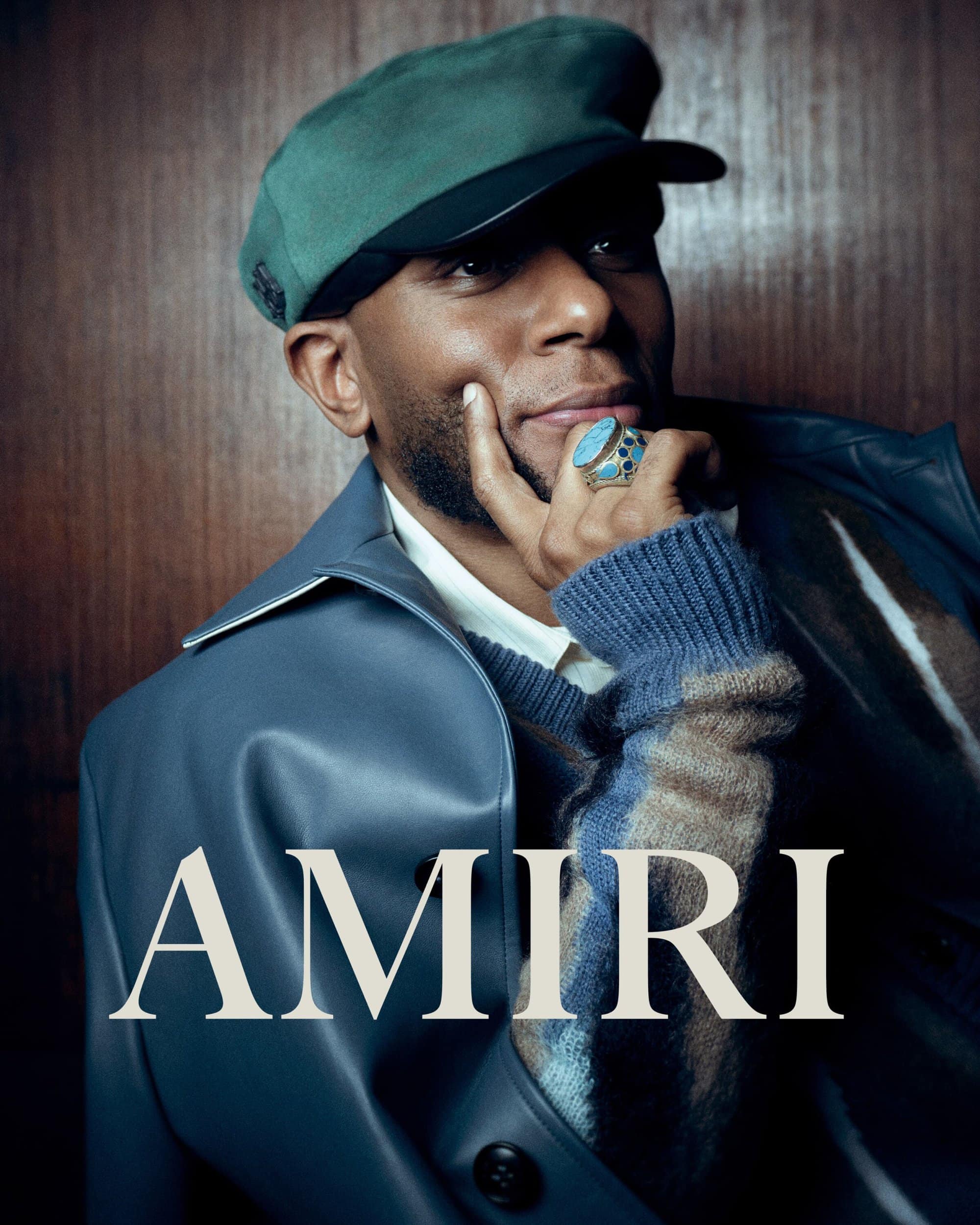 Portrait of a stylish adult male wearing a green cap, blue jacket, and statement ring, posing against a wood-panelled wall in studio lighting.