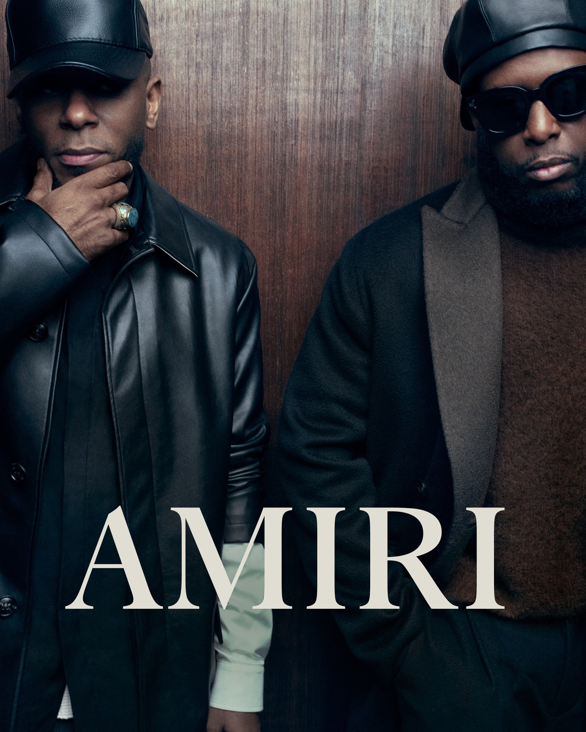 Two adult Black men pose indoors against a wood panel backdrop. Both are wearing jackets and hats; "AMIRI" text overlay.