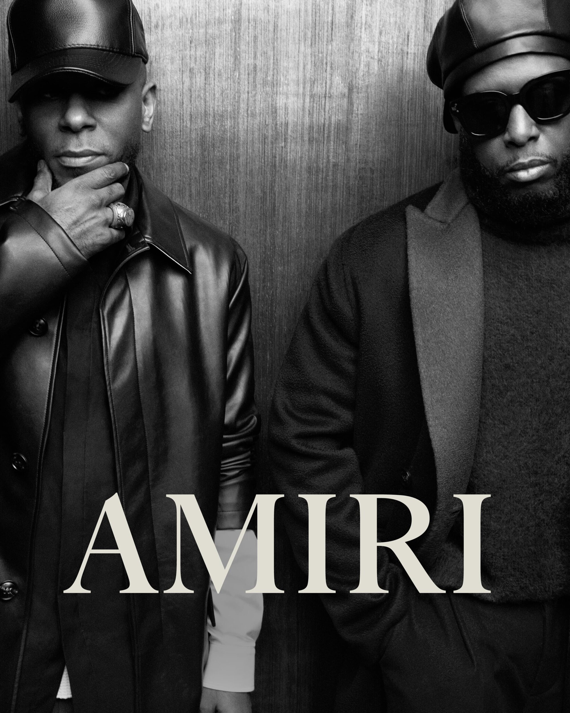 Two adult males stand indoors against a neutral wall, both wearing hats and dark jackets, with the word 'AMIRI' displayed.