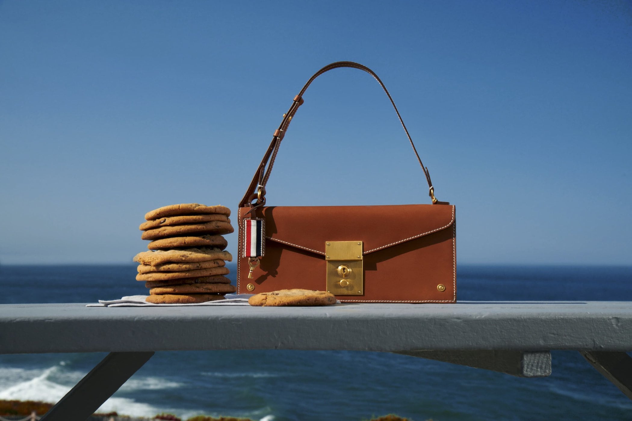 Luxury handbag paired with stack of cookies on a table against the backdrop of the ocean and clear blue sky. Food and fashion concept.
