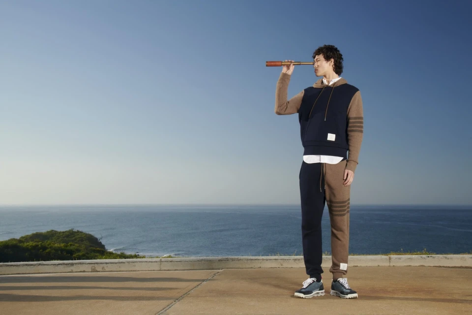 Man in stylish sportswear stands outdoors against ocean backdrop, looking through telescope, evoking a sense of exploration and fashion.