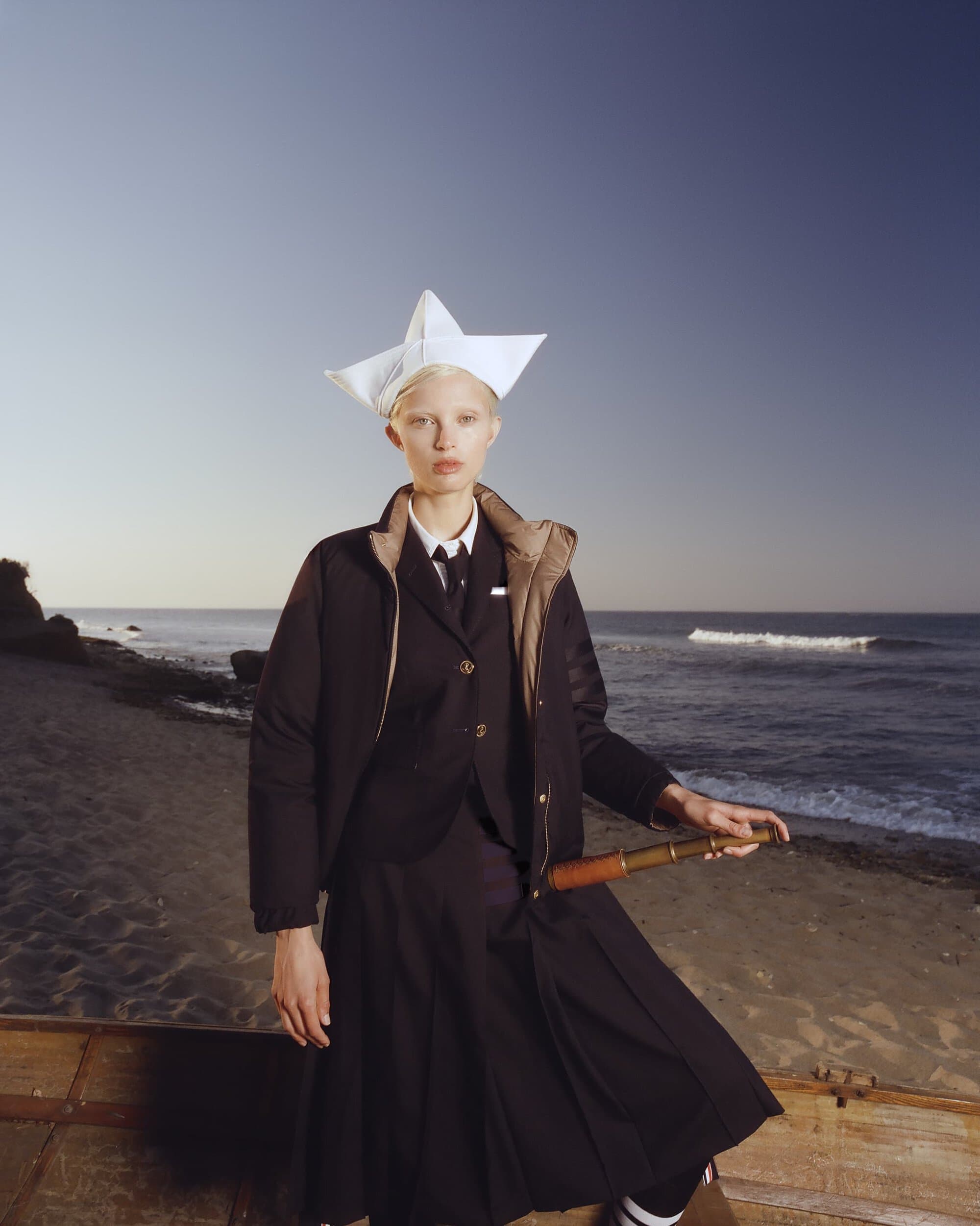Fashion portrait on a beach featuring an adult female wearing a dark suit, a paper hat, and holding a vintage telescope.