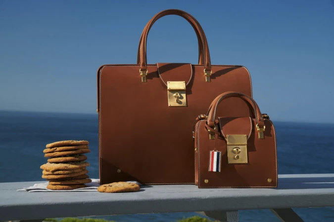 Luxury fashion: a large and small brown leather handbag sit beside a stack of cookies with an ocean backdrop under a clear blue sky.