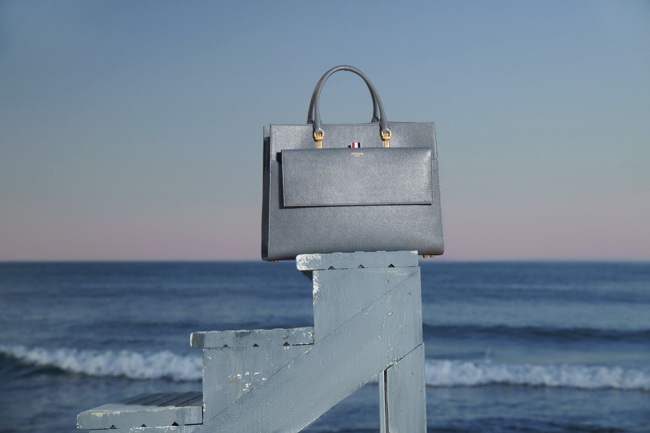 Grey leather tote bag with top handle and front flap pocket sitting on top of blue painted lifeguard stand with ocean and sky background.