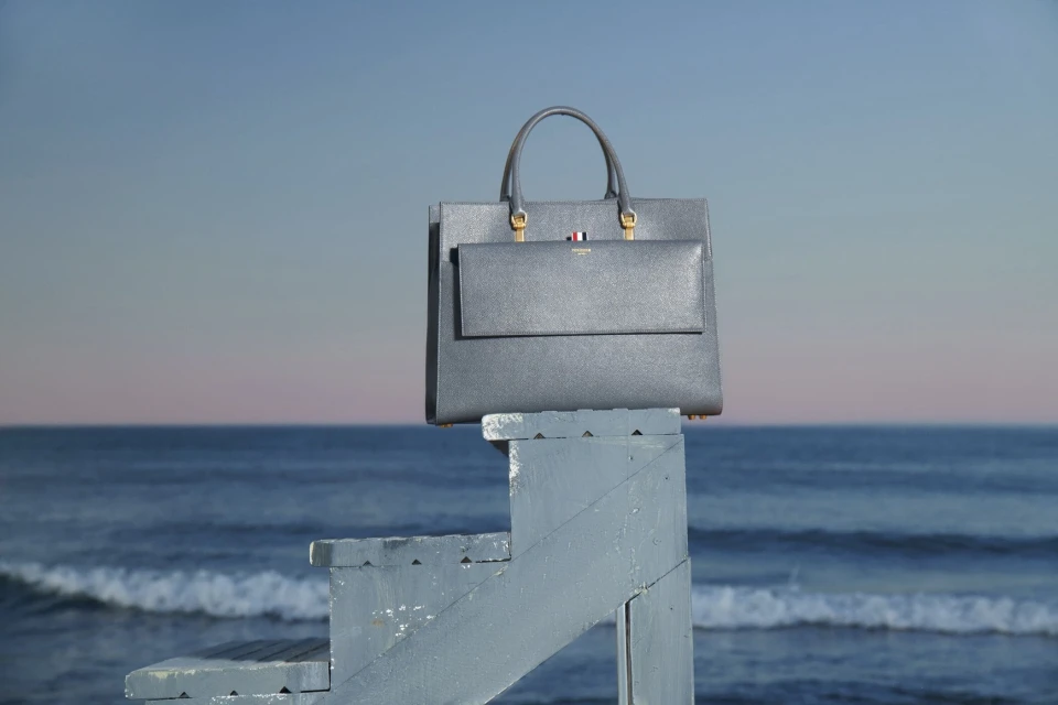 Grey leather tote bag with top handle and front flap pocket sitting on top of blue painted lifeguard stand with ocean and sky background.